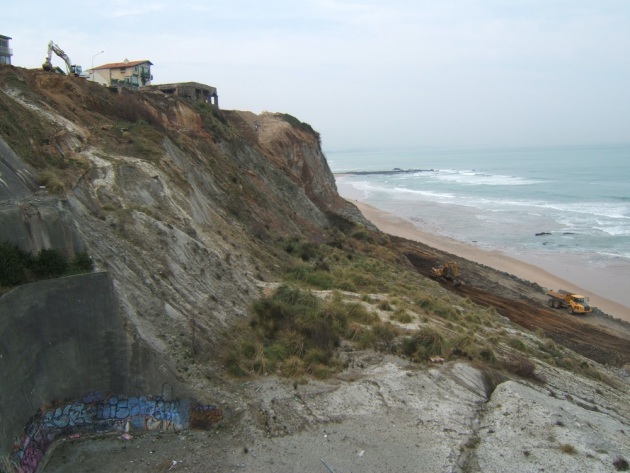 Photo d'une falaise, une grue est visible au sommet, des maisons sont en cours de destruction. Des camions sont visibles sur la plage en contrebas.