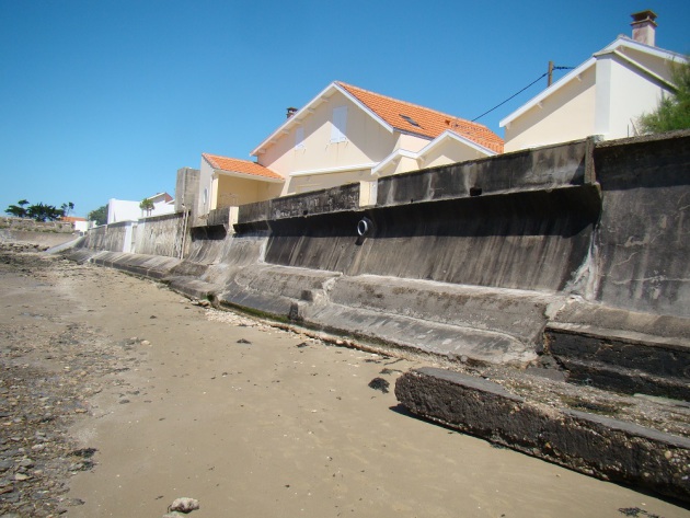 Photo prise depuis la plage, des infrastructures en béton sont présentes devant les maisons, un affaissement du sable devant le béton commence a être visible.