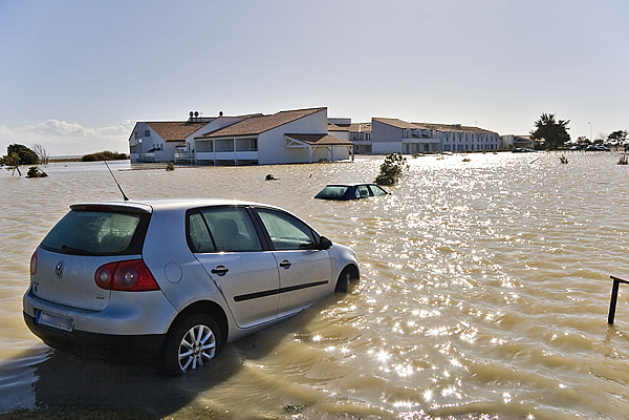 Suite à la tempête, une voiture au premier plan à les roues dans l'eau trouble, au second plan on aperçois le toit d'un autre véhicule et à l'arrière plan des bâtiments entourés d'eau