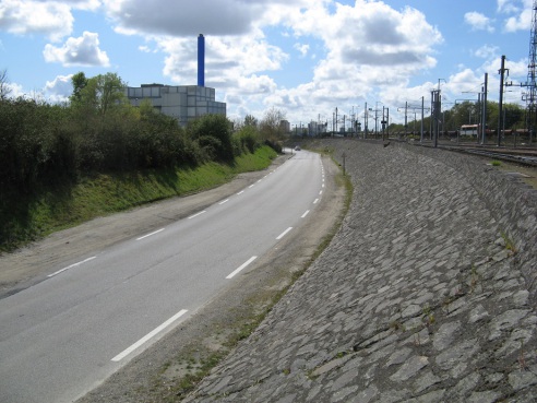 Route en aplomb d'une ligne de chemin de fer, on peut voir le toit d'un bâtiment industriel.